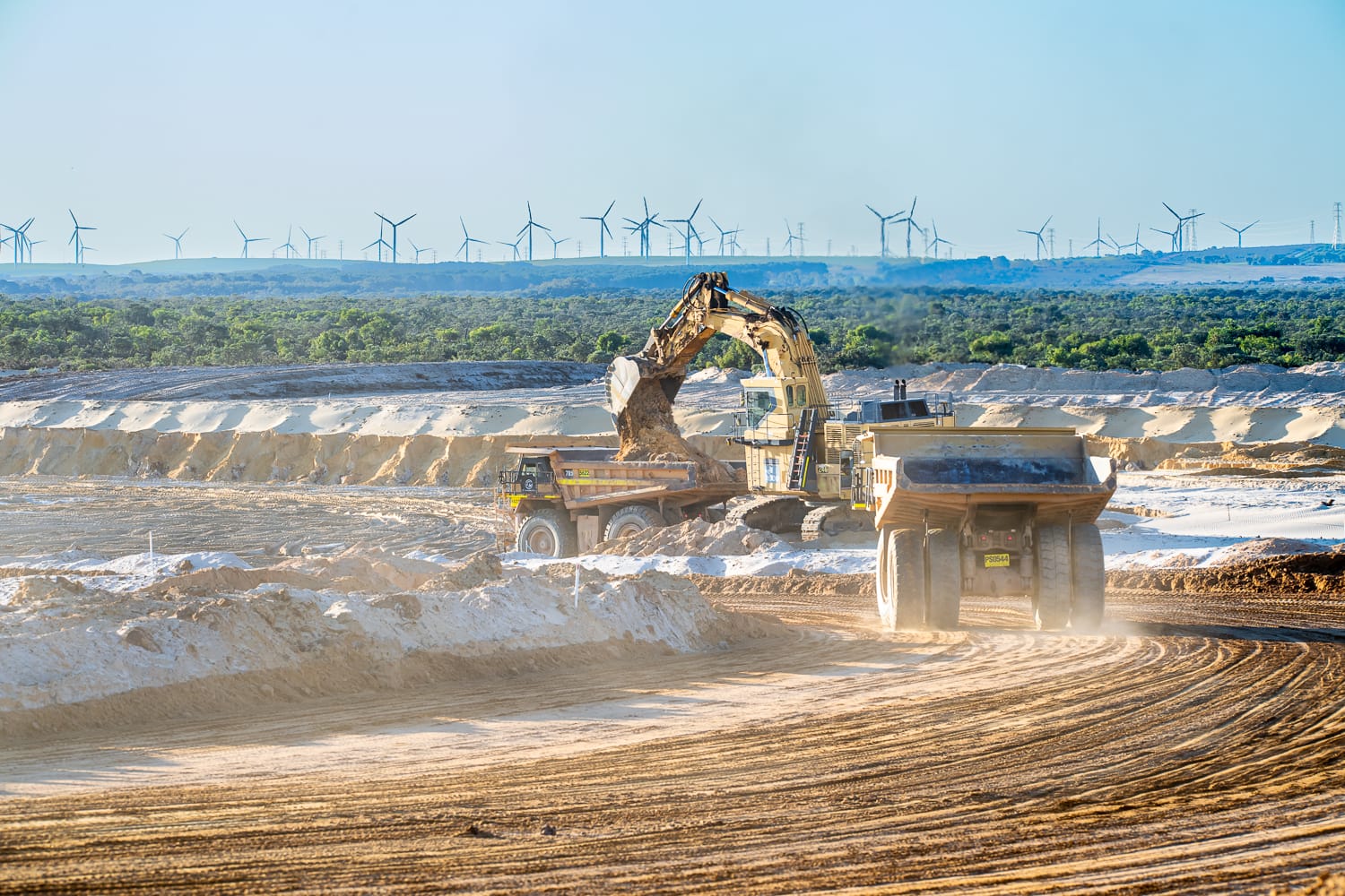 Construction site with excavators and dump trucks moving earth. Wind turbines are visible in the background under a clear blue sky.