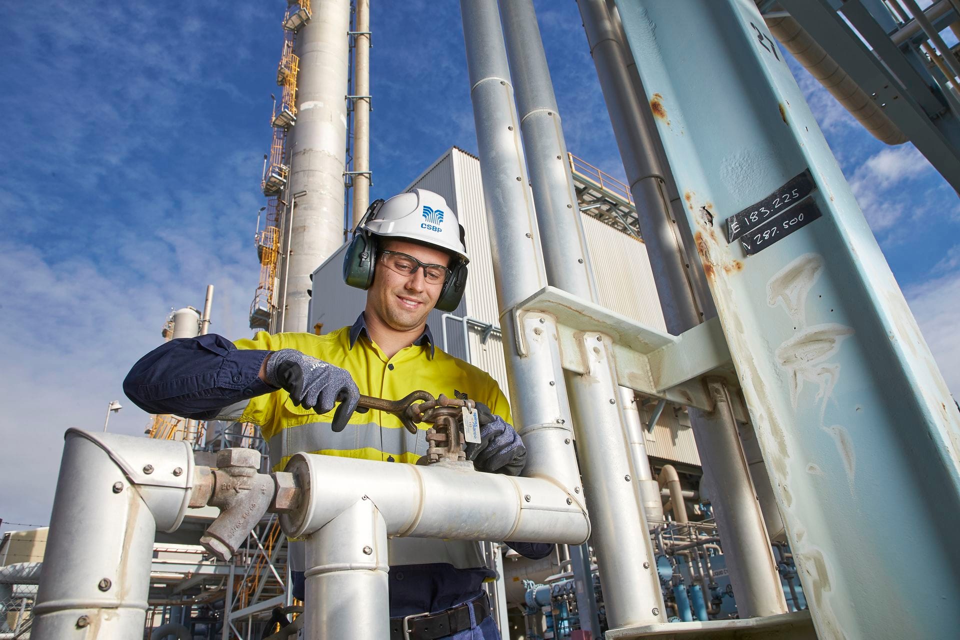 Worker in safety gear operates machinery at an industrial site with pipes and tanks under a blue sky.